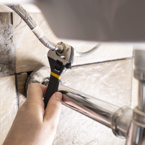 plumber hand fixes flexible hoses under sink by adjustable wrench closeup at home